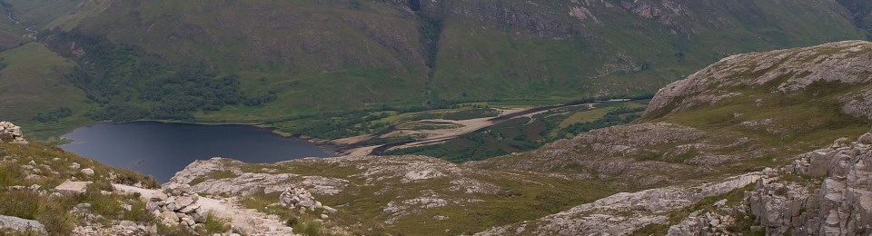 Dog on rocky trail peering down the hill toward a tarm with mountains in background