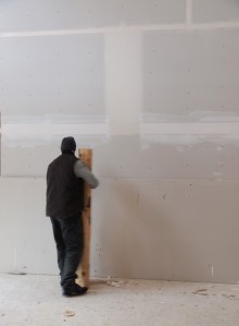 Man holding half of a 48" log that will become a stair silhouetted against blank dry wall that has been taped but not textured