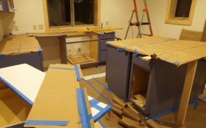 kitchen under construction showing counters covered by brown craft paper with tile designs hand drawn on paper. Room is messy with scraps of paper and tile templates. Ladder shows in back