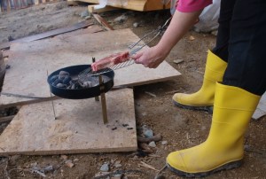 tiny charcole grill sitting on three quarter inch plywood on ground with woman holding grill rack with huge steak. The woman has on yellow mud boots.